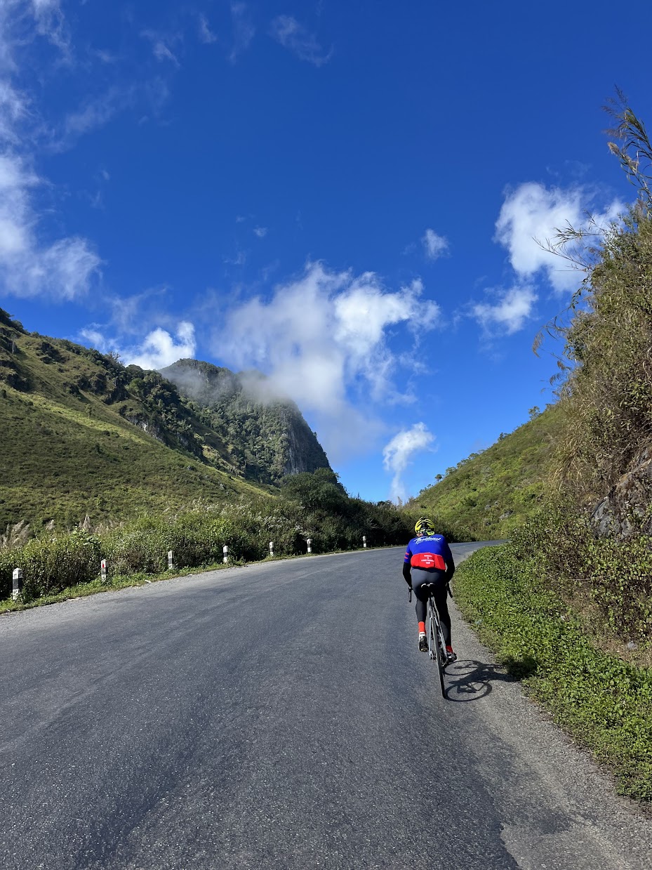 A cyclist climbing a paved road heading toward lush green mountains under a blue sky on our guided Laos bike tour.