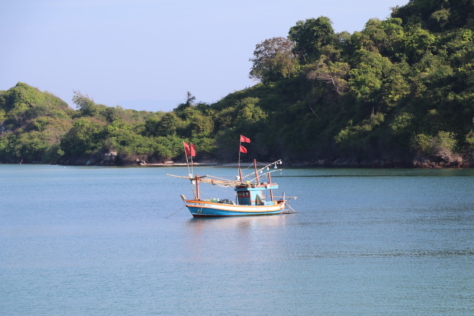 A traditional wooden fishing boat floating on calm turquoise waters near a forested island shoreline on our coastal cycling tours.
