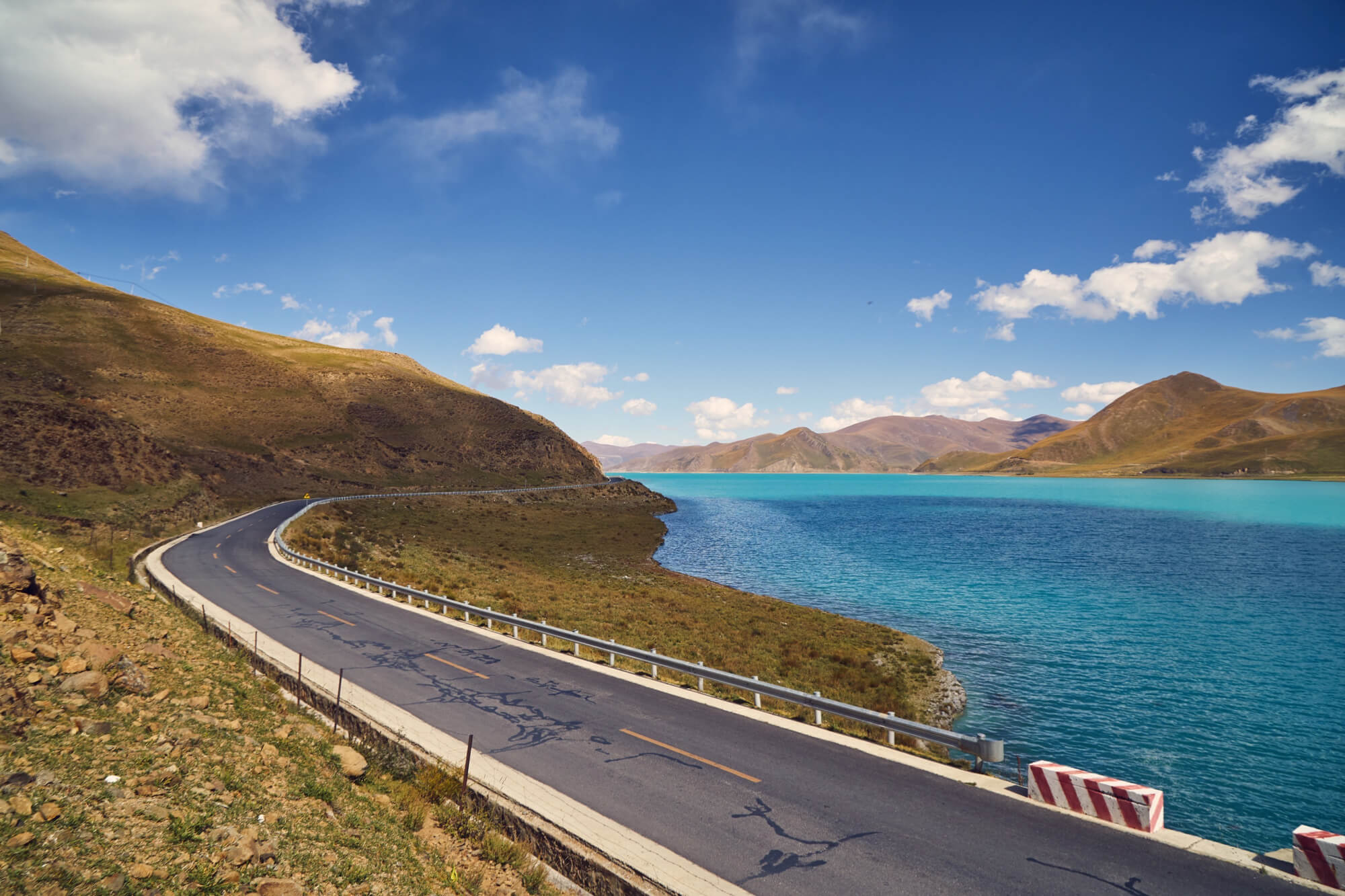 A winding asphalt road running next to the bright turquoise waters of Yamdrok Lake on our Epic Tibet road cycling expedition.
