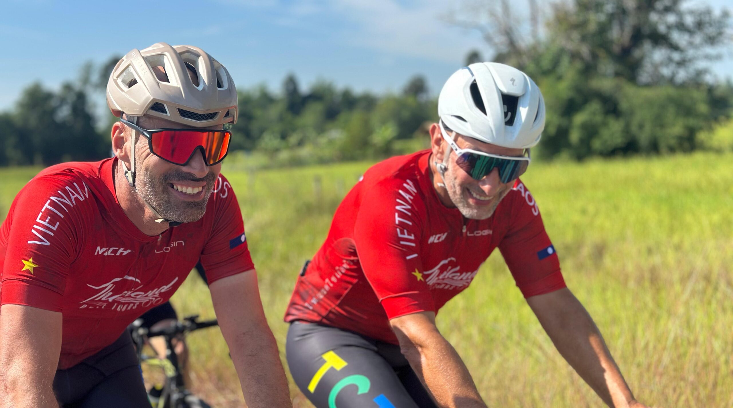 Two road cyclists smiling and riding side-by-side in matching team kits on a fully supported long-distance cycling holiday.