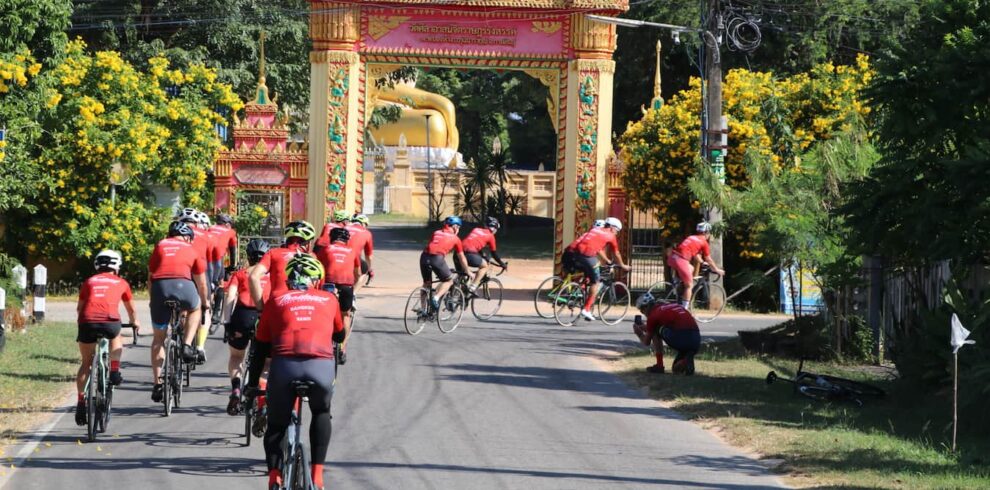 Cyclists on a road bike tour riding through an ornate traditional golden gate of a Buddhist temple in rural Thailand.