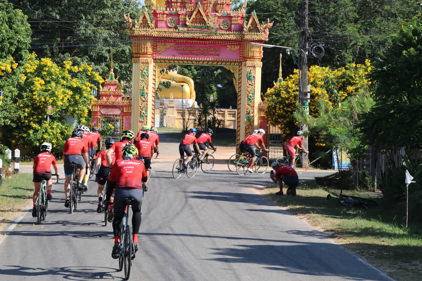 Cyclists on a road bike tour riding through an ornate traditional golden gate of a Buddhist temple in rural Thailand.
