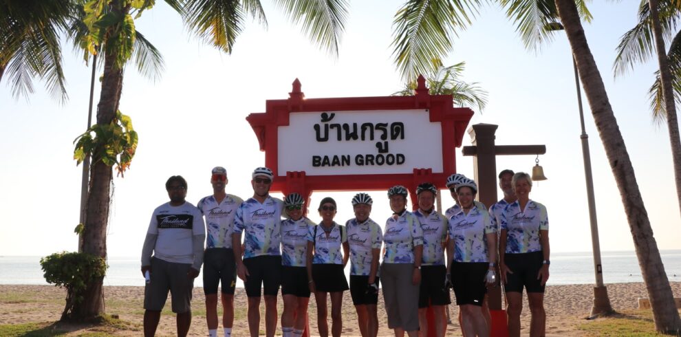 Cyclists in team kits posing under the traditional wooden Baan Grood welcome sign on the beach in Ban Krut.