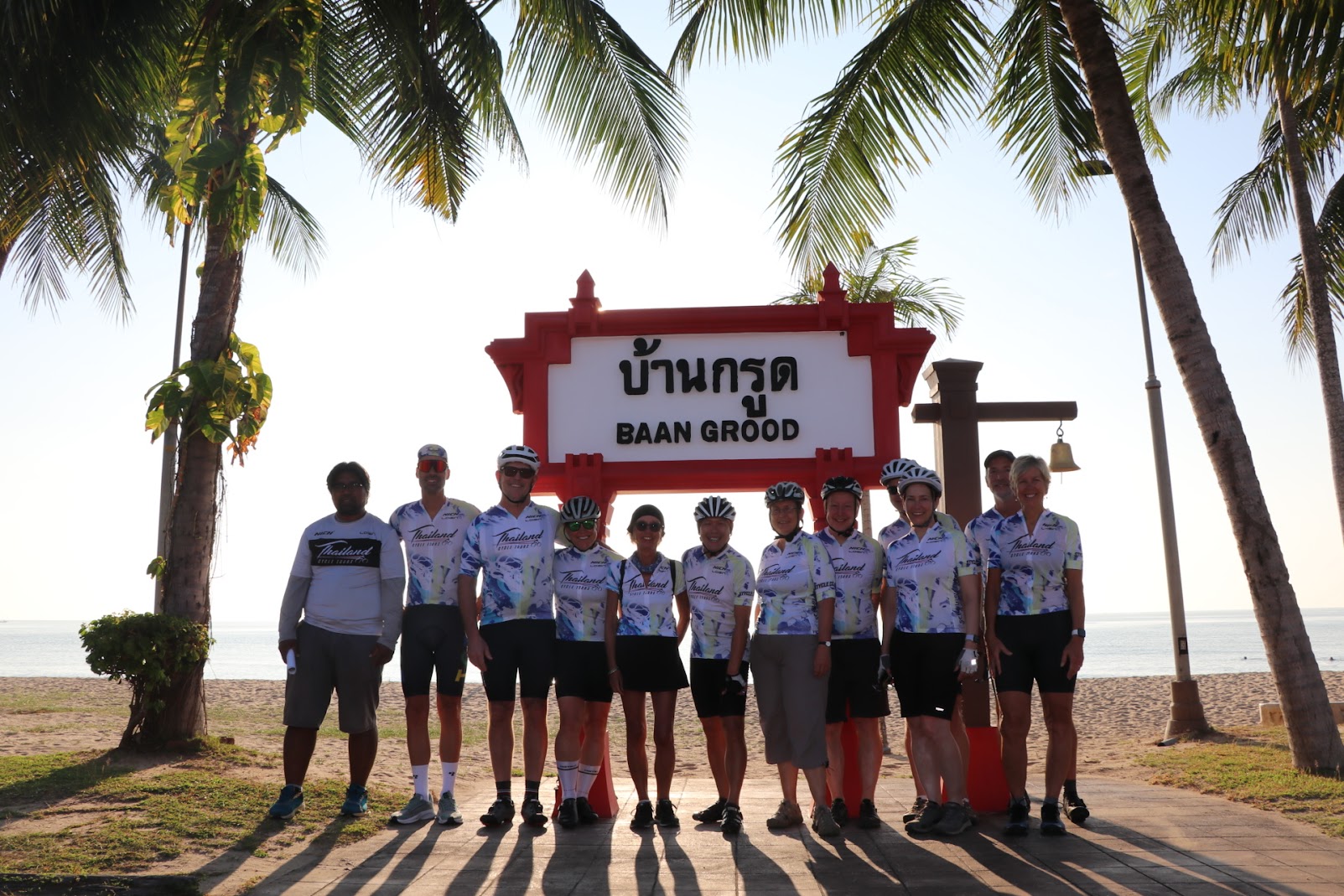 Cyclists in team kits posing under the traditional wooden Baan Grood welcome sign on the beach in Ban Krut.