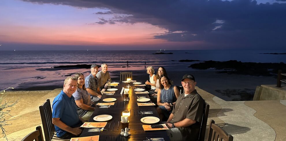 A group of cyclists enjoying an open-air seafood dinner at a long table on the beach during sunset.
