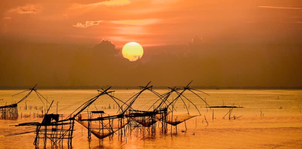 A breathtaking golden sunset over Patthalung Lake, showing traditional giant bamboo dip nets during a scenic boat tour.