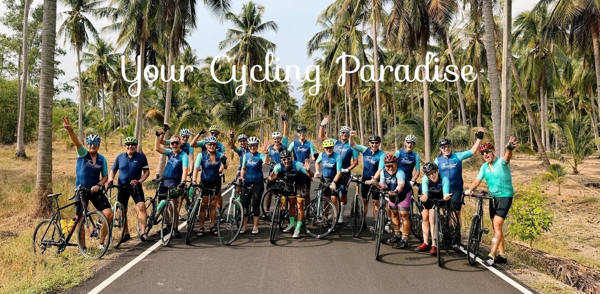 A large group of cyclists posing with their road bikes on a quiet, palm-lined lane in tropical Thailand.