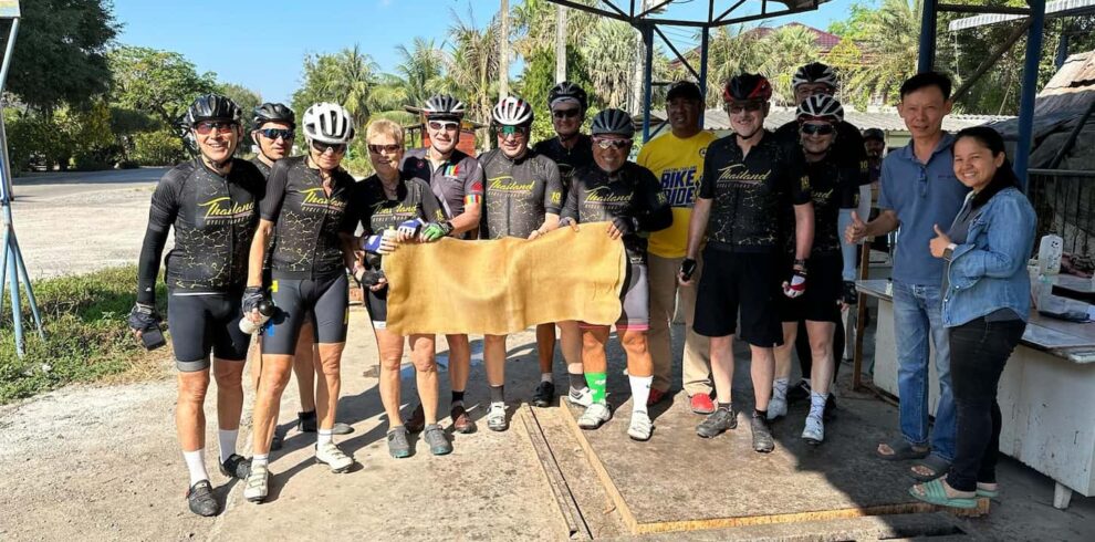 Cyclists on a fully supported bike tour in Thailand posing with a raw rubber sheet at a local plantation stop.
