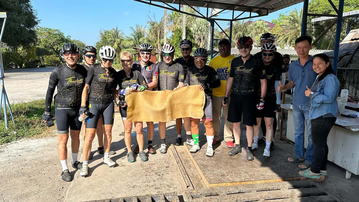 Cyclists on a fully supported bike tour in Thailand posing with a raw rubber sheet at a local plantation stop.