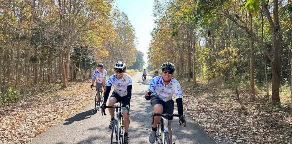 Cyclists in matching team jerseys riding on a tree-lined back road during a guided cycling tour in Thailand.