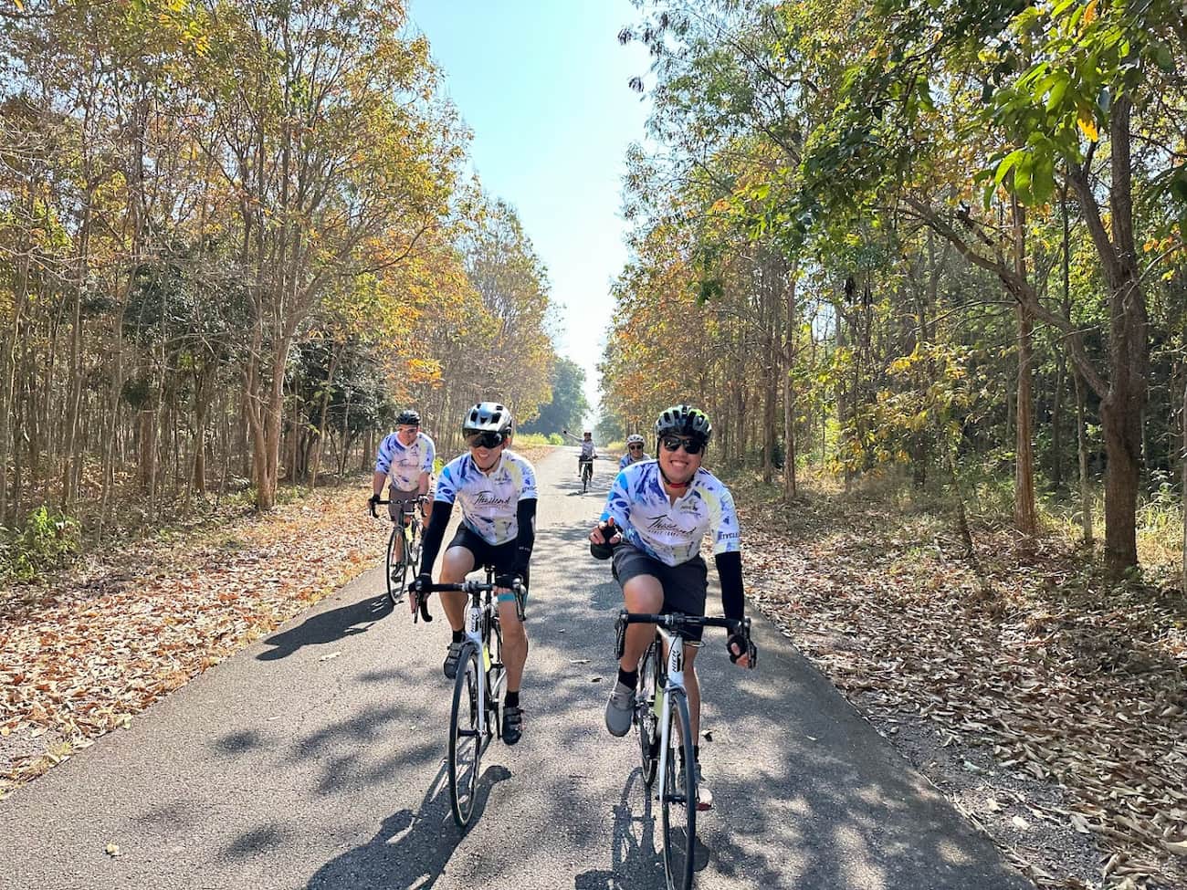 Cyclists in matching team jerseys riding on a tree-lined back road during a guided cycling tour in Thailand.