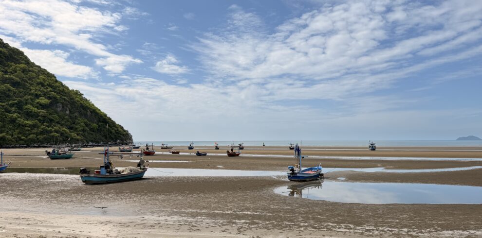 Traditional fishing boats resting on tidal sand flats along a peaceful coastal bay, surrounded by forested hills and expansive sea views under a bright blue sky in southern Thailand.