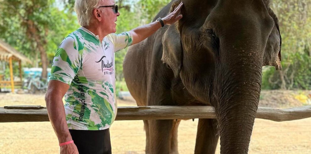 A senior road cyclist wearing a green and white team kit interacting with an elephant during an elephant safari excursion.