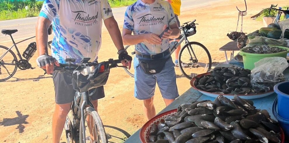 Tour cyclists visiting a traditional roadside seafood stall, discovering local produce and everyday life along a scenic cycling route through southern Thailand.