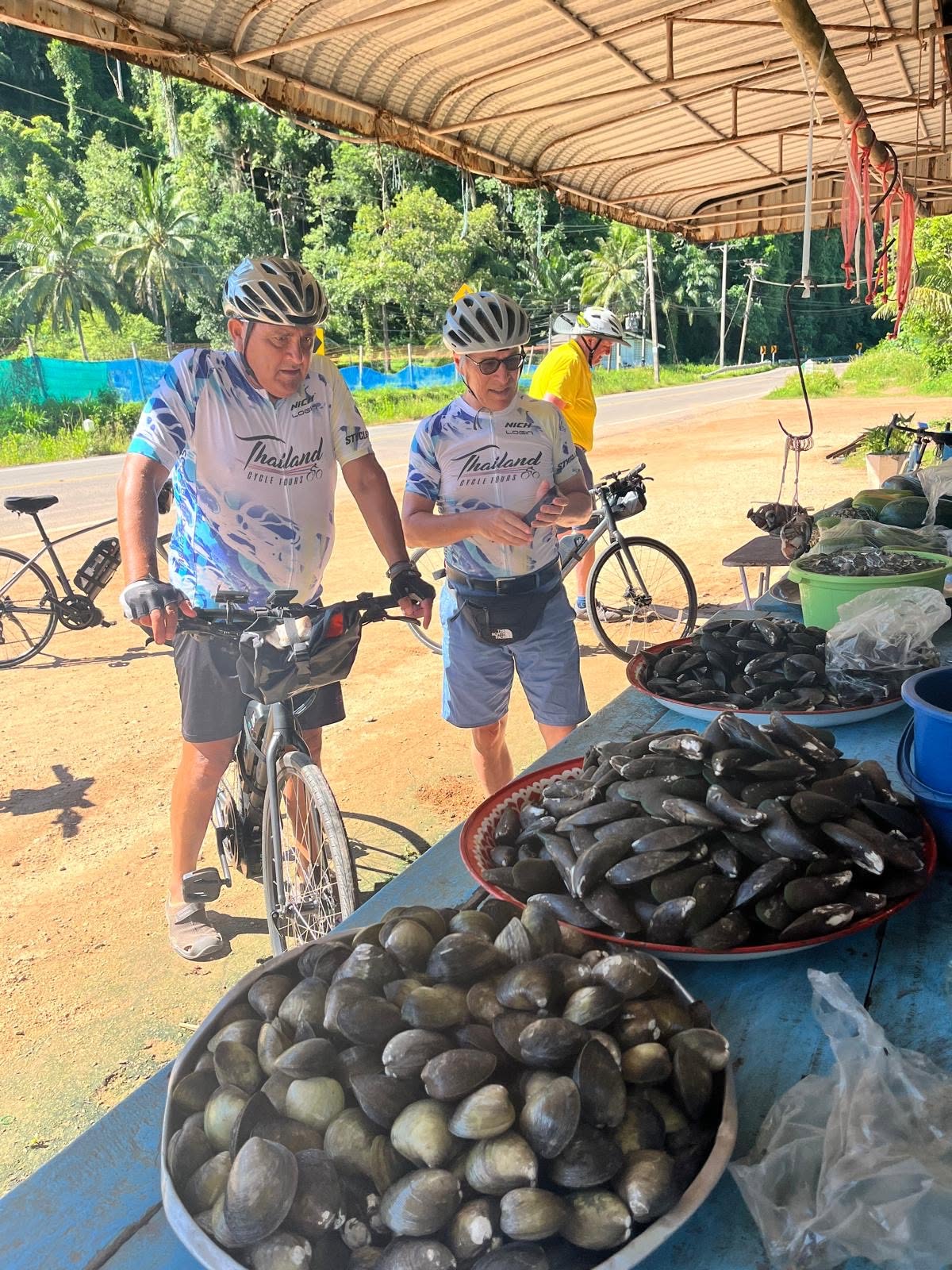 Tour cyclists visiting a traditional roadside seafood stall, discovering local produce and everyday life along a scenic cycling route through southern Thailand.