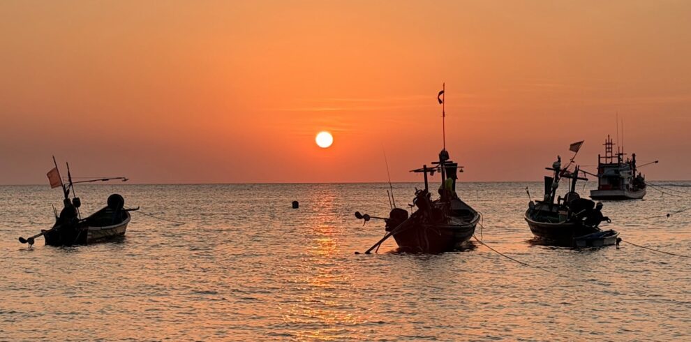 Traditional Thai longtail fishing boats anchored on calm sea waters during a vibrant sunset, with the orange sun reflecting across the Gulf of Thailand. A peaceful coastal scene showcasing local fishing culture, tropical seascapes, and the natural beauty of Southern Thailand.