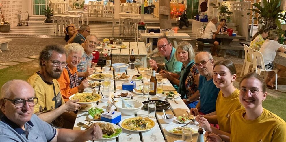 Group of cyclists and travelers enjoying a traditional Thai dinner at an outdoor restaurant, gathered around a communal table under warm string lights during a cycling tour in Thailand.