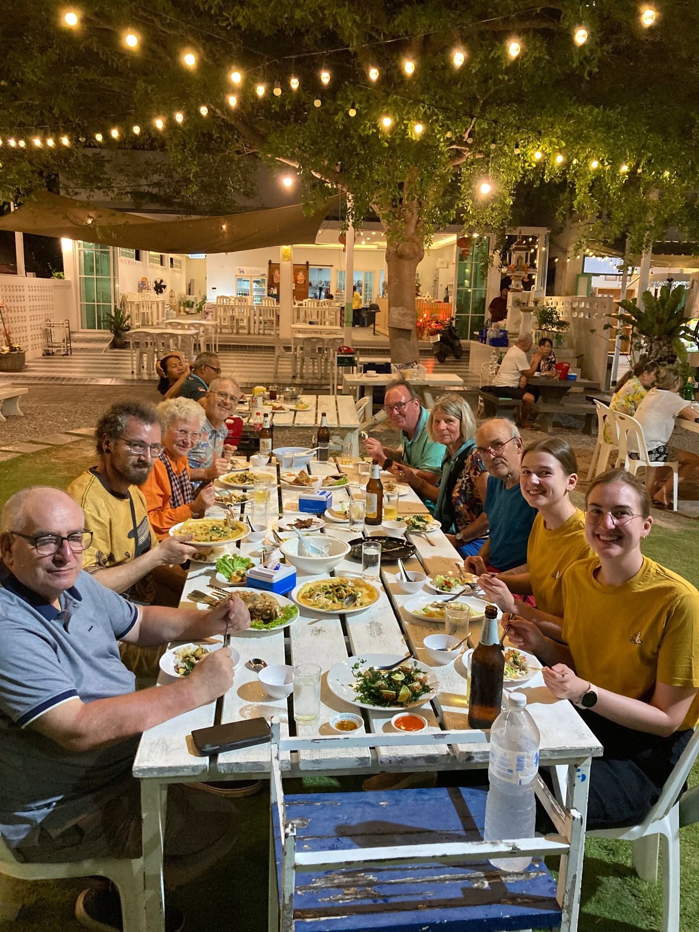 Group of cyclists and travelers enjoying a traditional Thai dinner at an outdoor restaurant, gathered around a communal table under warm string lights during a cycling tour in Thailand.