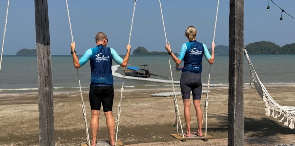Two cyclists in cycling gear relaxing on a swing overlooking the stunning coastline of Sawi, Thailand. The scenic view features a pristine beach, turquoise ocean waters, lush national park landscapes, and tropical islands in the distance, showcasing the beauty of cycling adventures with Thailand Cycle Tours.