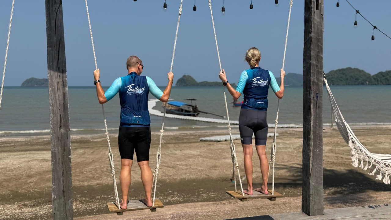 Two cyclists in cycling gear relaxing on a swing overlooking the stunning coastline of Sawi, Thailand. The scenic view features a pristine beach, turquoise ocean waters, lush national park landscapes, and tropical islands in the distance, showcasing the beauty of cycling adventures with Thailand Cycle Tours.