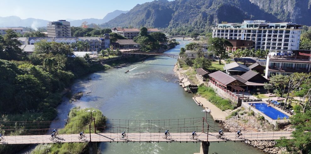 Cyclists crossing a suspension bridge in Vang Vieng, Laos, with the Nam Song River, limestone mountains, and riverside resorts in the background.