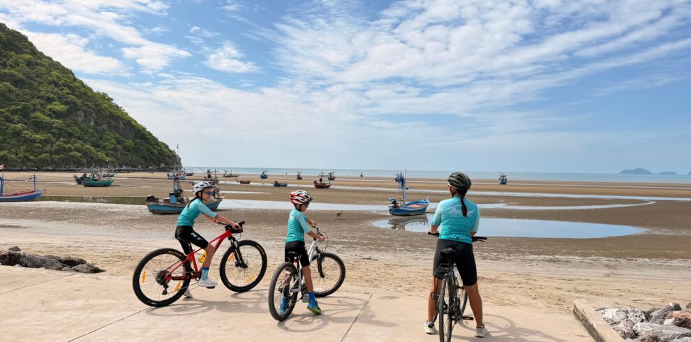 Family cycling tour in Thailand with parents and children riding bicycles along a scenic coastal road, surrounded by tropical beaches, palm trees, turquoise sea views, and lush green hills on a sunny day. Perfect for active family holidays with Thailand Cycle Tours.