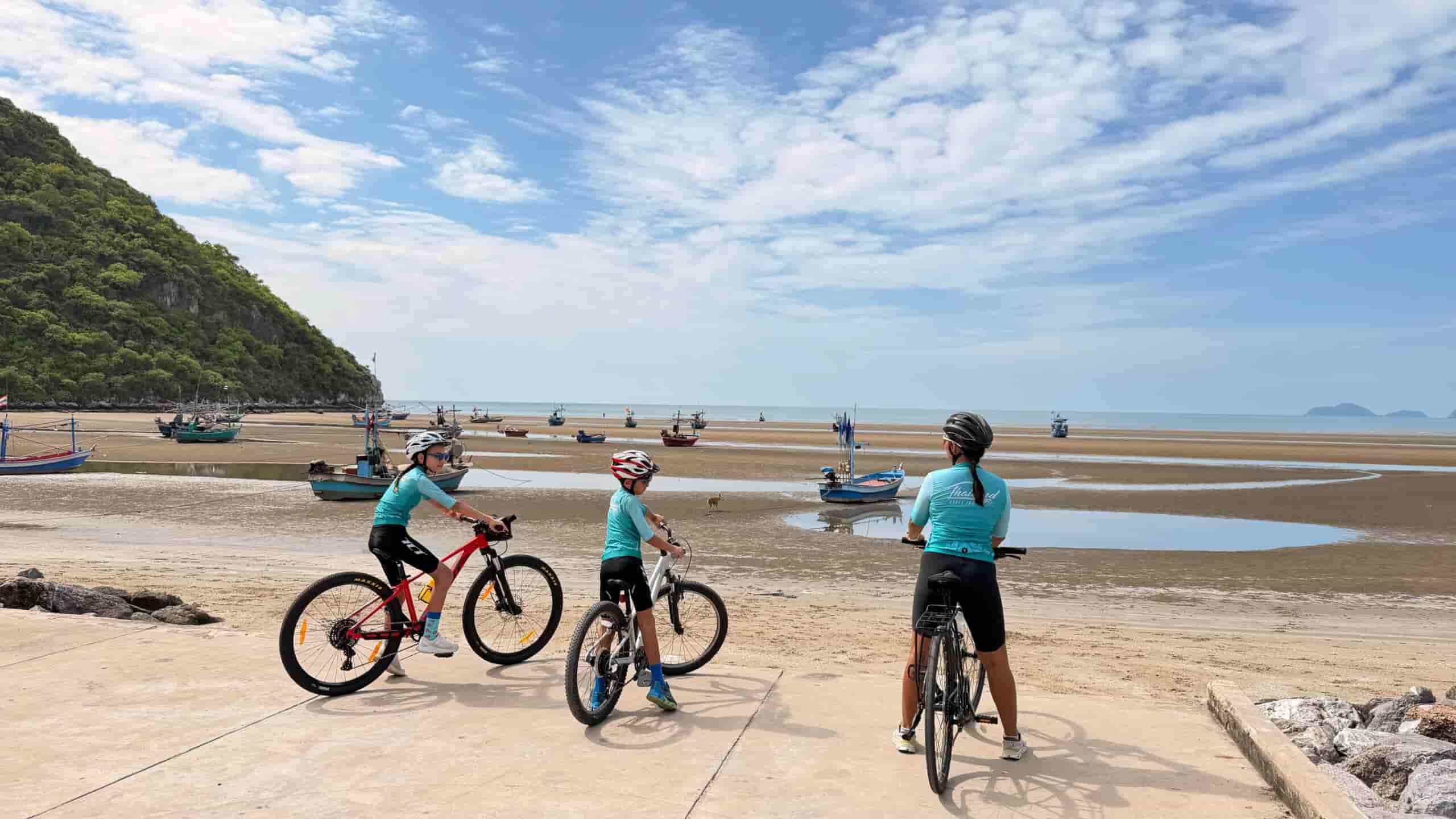 Family cycling tour in Thailand with parents and children riding bicycles along a scenic coastal road, surrounded by tropical beaches, palm trees, turquoise sea views, and lush green hills on a sunny day. Perfect for active family holidays with Thailand Cycle Tours.