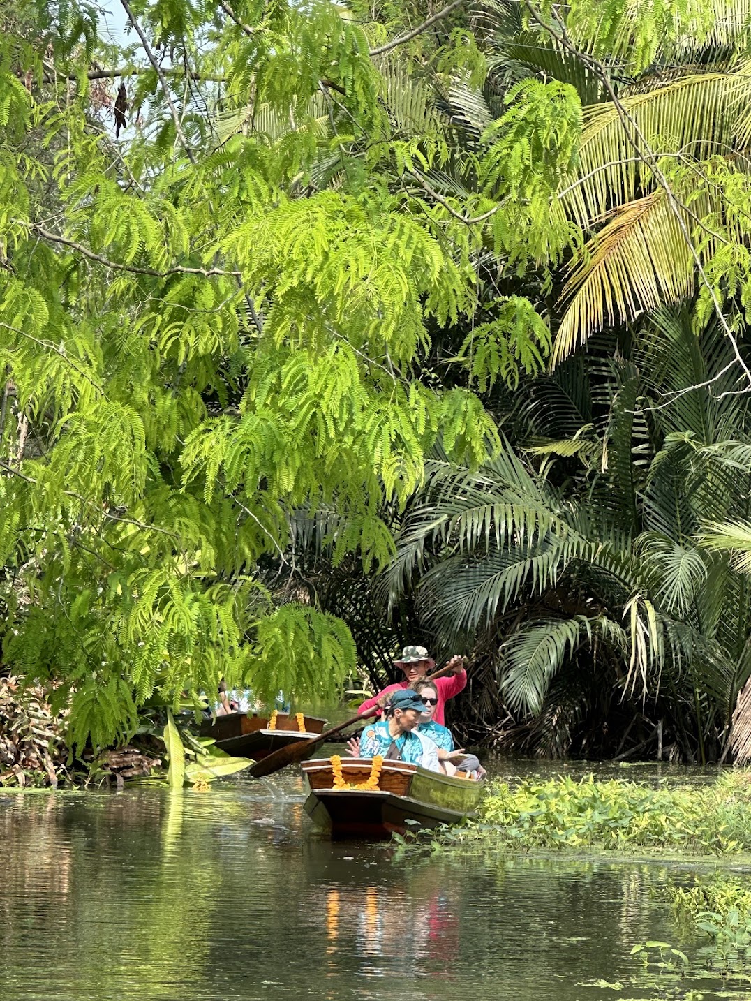 Traditional wooden boat gliding through a lush tropical canal, surrounded by dense palm forests and vibrant greenery, offering an authentic river journey through Thailand’s tranquil waterways.