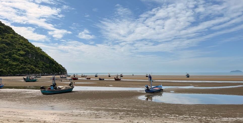 Fishing boats resting on the sand flats of Khao Kalok Bay at low tide, with scenic coastal views and blue skies along the Thailand Cycle Tours cycling route near Pranburi, Thailand.