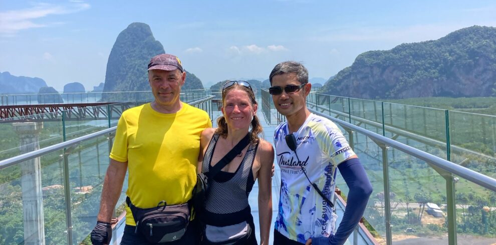 Cyclists posing on a glass skywalk with breathtaking views of limestone cliffs, tropical forests, and coastal scenery in southern Thailand.