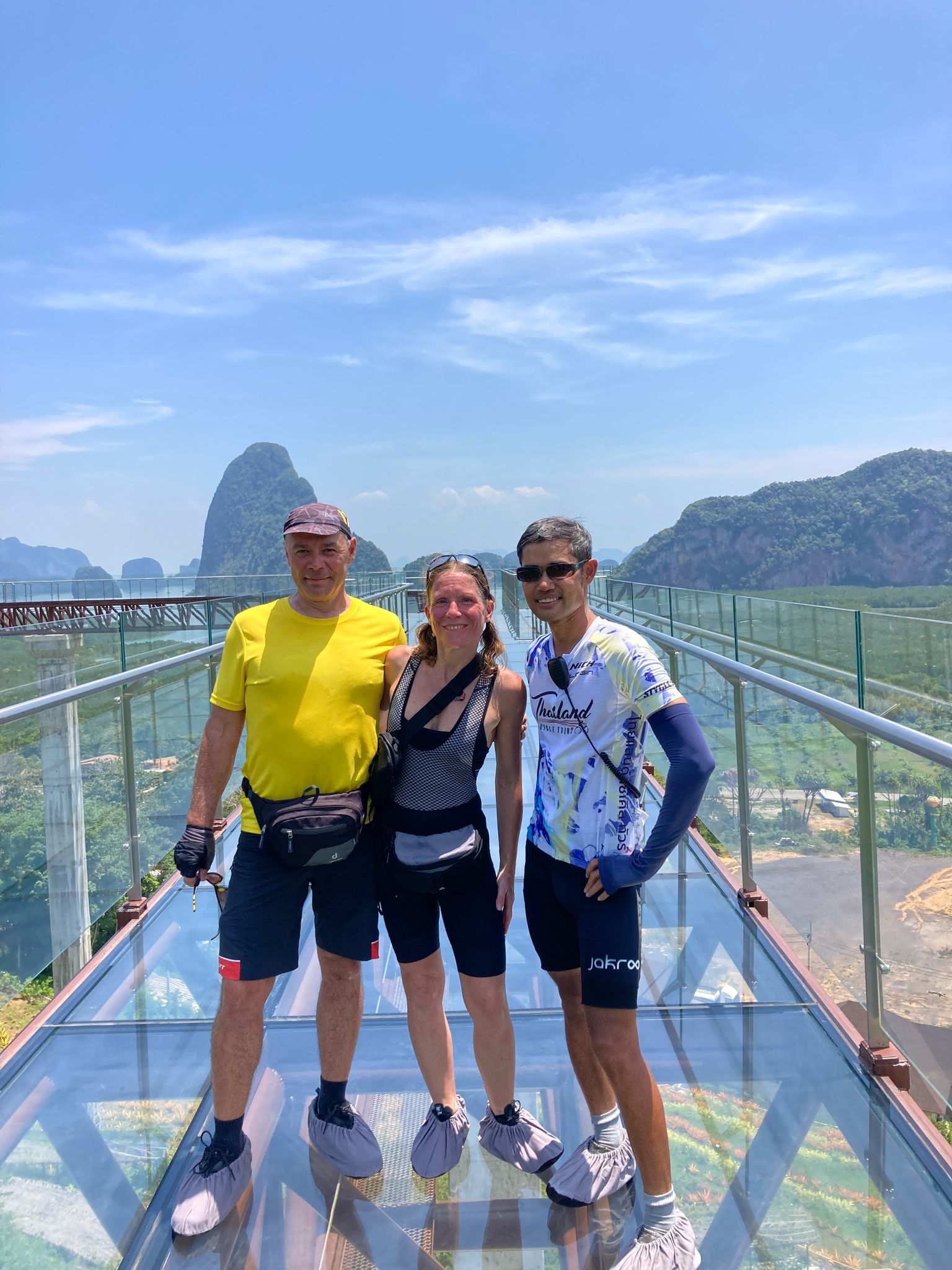 Cyclists posing on a glass skywalk with breathtaking views of limestone cliffs, tropical forests, and coastal scenery in southern Thailand.