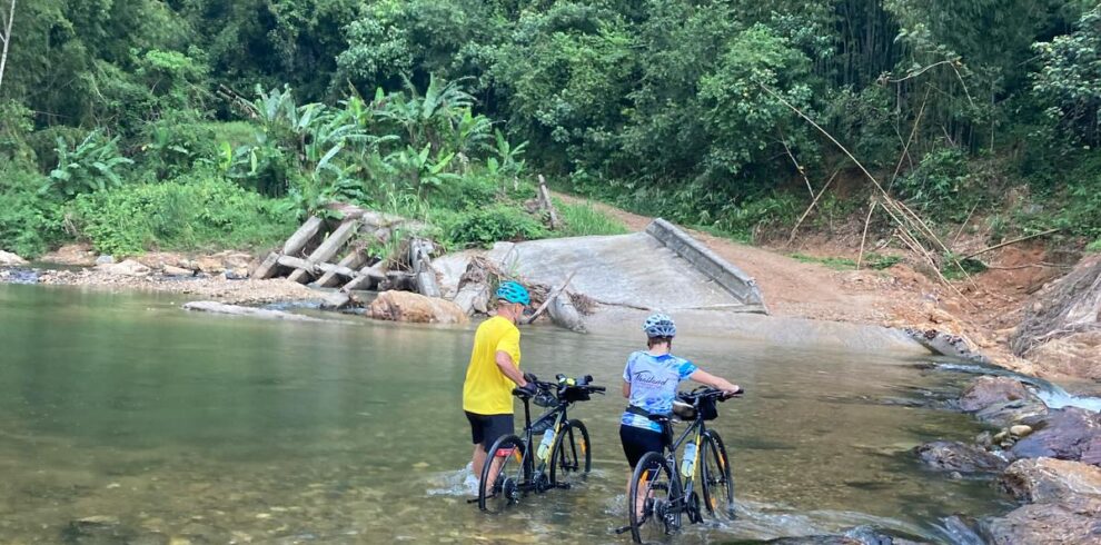Two cyclists walk their bikes across a shallow river surrounded by lush tropical forest.