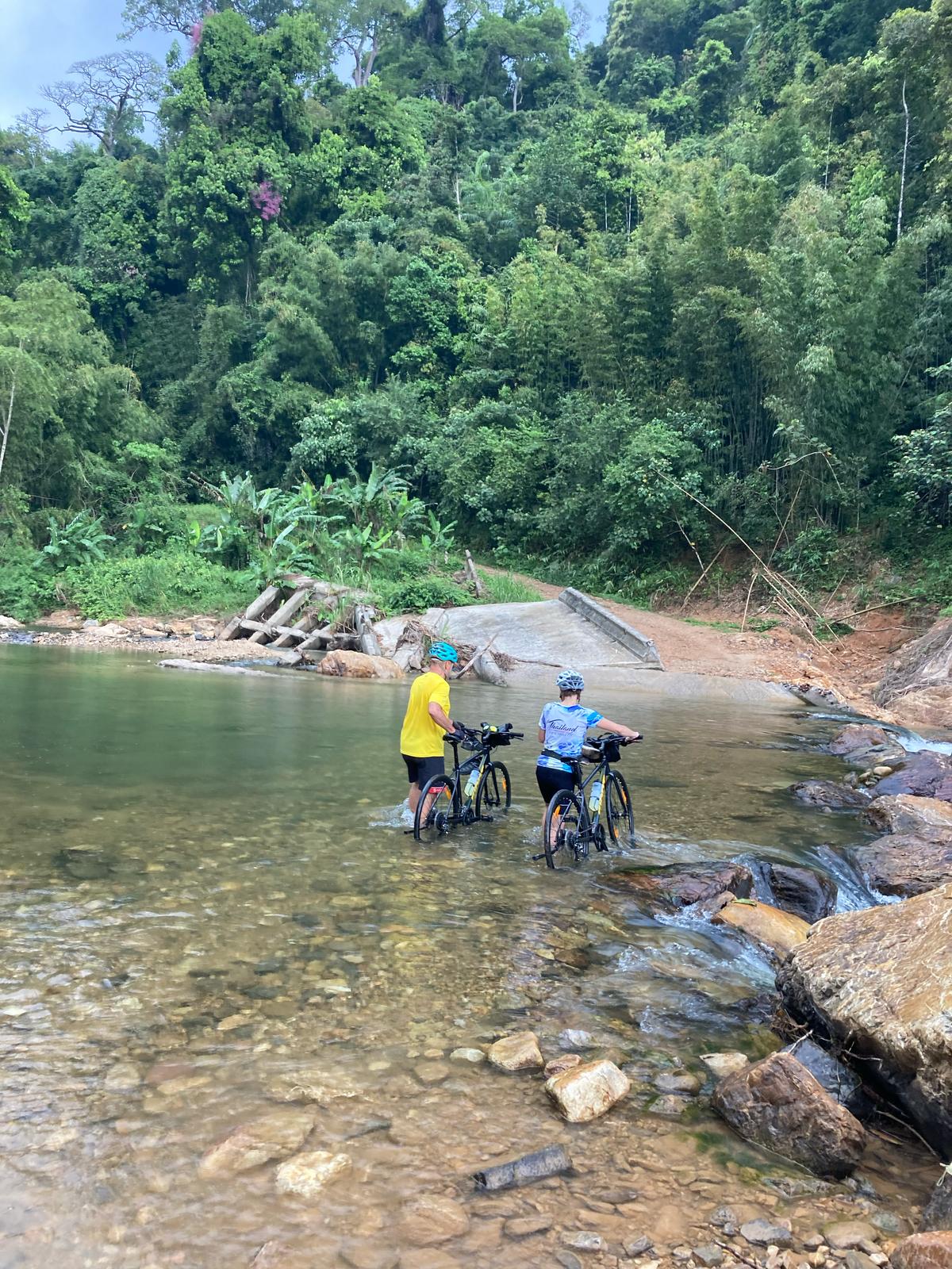 Two cyclists walk their bikes across a shallow river surrounded by lush tropical forest.