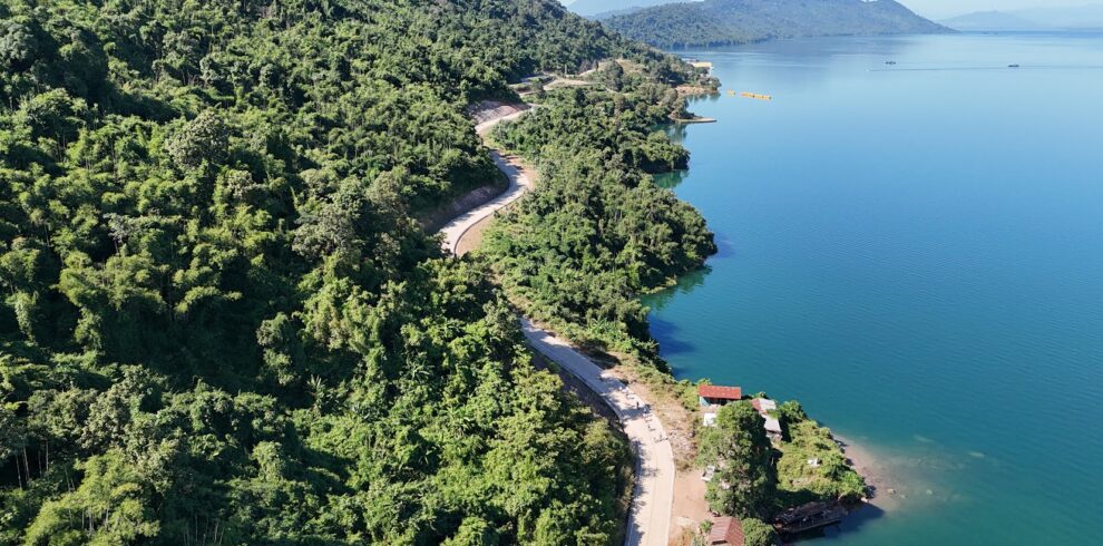 Cyclists riding on a beautiful scenic road along Nam Ngum Lake in Laos, surrounded by tranquil blue water, rolling hills, and lush tropical landscapes during a cycling tour with Thailand Cycle Tours.
