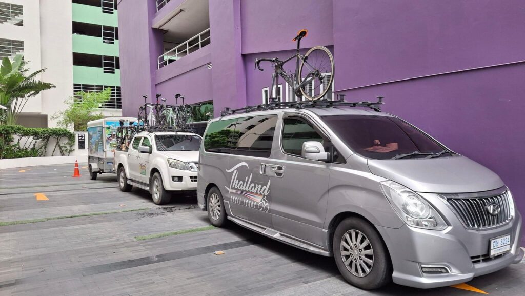 Thailand Cycle Tours support van and pickup truck with bicycles mounted on roof racks, parked outside a building.